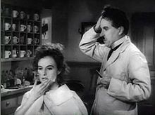 A black-and-white screenshot of a woman sitting in a chair. She is rubbing her face and a barber is standing at her right and is holding his head with his right hand. The two people are located in a barbershop.