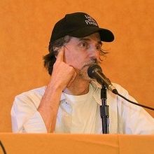 upper body shot of man sitting at desk with white shirt and black baseball cap speaking into microphone