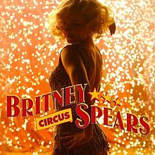 The torso of a young blond woman. She has a wavy blond hair cut and is wearing a sparkly golden dress. She is looking over her shoulder. Her left hand is resting on her left hip. In the back, there is a shower of sparks. The words "BRITNEY" followed by three yellow stars and "SPEARS" are written in capital red circus-like handwriting. Below "BRITNEY", the word "CIRCUS" is written in smaller capital letters.