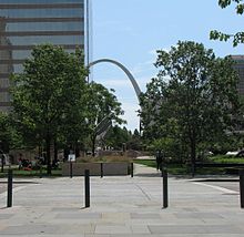 Four black bollards sit in the foreground, trees are visible in the middle, and in the distant background, a tall grey arch peeks out from behind office buildings.