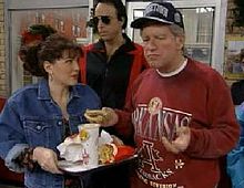 A man stands on the right dressed in a baseball cap and sweatshirt to resemble President Clinton. He is holding a burger which he has picked up from the women to his left's tray; several other products remain. A man in dark glasses stands behind them.