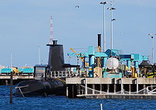 A submarine next to a dock, which is occupied by several cranes and other mechanical equipment