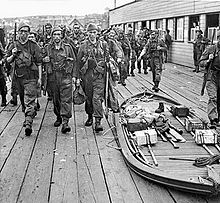 Armed soldiers march past a collapsed boat containing equipment
