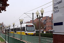 A white and yellow train arriving at a station with several lamp posts in the foreground, transit oriented development at the rear and station signage at the far right.
