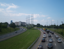 A six lane freeway curving to the right. The opposing directions of travel are separated by a grassy median. Power lines cross the highway, and apartments are visible in the background