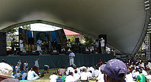 Performers on an outdoor stage with shell-shaped overhang before an audience seated in the sun.