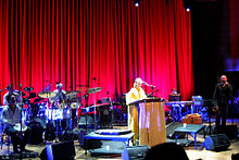 An ensemble are performing on-stage, three musicians are seated at extreme left behind musical instruments. Lisa Gerrard is behind a lectern near mid-stage with a microphone. At the right is Brendan Perry holding a microphone with his left hand. His right hand is alongside his thigh and holding an instrument. The background includes a long stage curtain with another musician seated at rear right, who is obscured behind a keyboard.