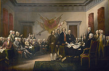 About 50 men, most of them seated, are in a large meeting room. Most are focused on the five men standing in the center of the room. The tallest of the five is laying a document on a table.