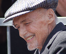 A smiling Dennis Hopper, appearing frail and aged, wearing a gray hat over his thin gray hair, with a bandage above his right eye.