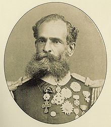 A photographic portrait of a dark-bearded man in heavily braided military tunic bedecked with many medals.