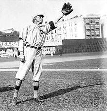 A man wearing a baseball uniform bearing the letter "P" on the chest leans slightly to his left and raises his left hand on which he is wearing a baseball glove.