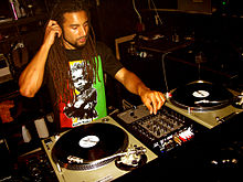Man sitting in a studio before two turntables and audio turning buttons, adjusting the sound as he listens over earphones.