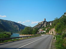 A view of a two lane road taken from a car. The road approaches a sharp left bend and a town, with a steep hill on the right and a river on its left.