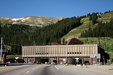 Automobiles are driving on a road leading to one of two openings in a building against a mountain. Letters above each opening read "Johnson Tunnel 1979" and "Eisenhower Tunnel 1973". On the roof of the building, large ventilation hoods are visible.