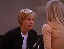 Still shot of a television show from 1997 showing, Ellen DeGeneres leaning forward to speak to a woman with long blond hair seen from behind. In between them is a microphone