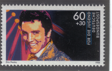 A horizontally formatted stamp with a close-up illustration of a young, smiling Presley. A bank of spotlights shines behind him.