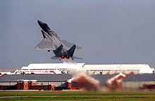 Gray jet fighter taking off at steep angle of attack, with full afterburner, as evident by hot gas ejected from its engines.