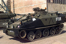 Spartan vehicle facing to the left, the driver can just be seen. To the rear is a Challenger 1 Main Battle Tank and a large concrete building