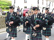 A group of men and women wearing traditional highland dress are playing the bagpipes as they walk down a road. A member of the group is playing a large red drum. A white, two storey building is in the background.