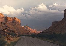 Highway through a sandstone gorge, with sunlight peering through clouds illuminating towers in the background.