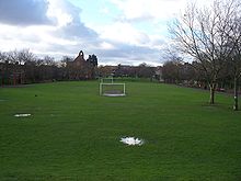 A long narrow strip of grass stretches into the distance, with houses on both sides. The grass is occupied by football pitches