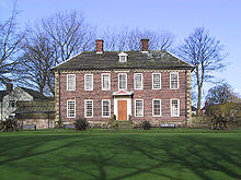 A two-storey brick-built mansion in its own well-kept garden of green grass. The ground floor has six windows - three on each side of a wooden front door. The second floor has seven windows. The house has a grey slate roof surmounted by two small brick chimneys. Behind the house are leafless trees and behind those, a clear blue sky.
