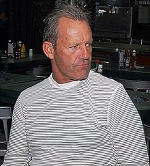 An older man with short gray hair in a black and white horizontally striped shirt looks to his left.