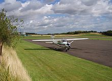 A tarmac runway is shown in the background. A small single engine plane is parked in the forecourt