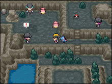 A pre-teenaged boy with black hair and a black and yellow baseball cap stands inside a dark, rocky, cave-like area. A small, blue, crocodile-like Pokémon stands behind him. Standing elsewhere in the area are two young men and one young woman, all wearing black clothes and beanies, and two small, pink, quadropedal Pokémon.