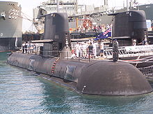 A submarine next to a dock, with navy personnel and civilians standing on the outer hull. Parts of another submarine and two warships can be seen in the background.