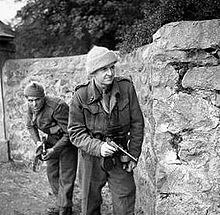 Two men in uniforms advance behind a wall carrying weapons
