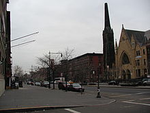 A two-lane street passes through a city neighborhood composed of a line of three-story buildings and a large church. The street is lined with decorative lampposts and small trees.