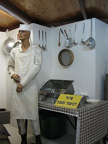 A museum mannequin standing in front of a table set with various kitchen utensils, with more kitchen utensils hanging from the wall behind