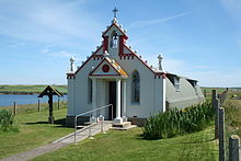 A small chapel sits in green fields under a blue sky. A body of water lies to the left. The front of the building is painted red and white and is decorated with colonnades and a small bell tower. By contrast the main part of the building is painted grey and has a curved exterior reminiscent of a Nissen hut.