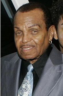 A mature African American man wearing a grey suit, black shirt and light blue tie. His hair is short and he wears a hooped earring in his right ear.