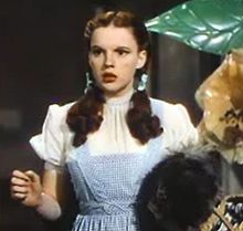 A color screenshot of a woman in blue overalls looking to her right while holding a dog in her left hand and raising her right to equal height.