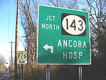 A green sign along a road reading junction north Route 143 Ancora Hospital. In the background to the left, a road approaching a traffic light is seen.