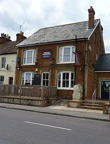 A photograph of a public house in a large red-bricked terrace house with a v-shaped slated roof, and an unpainted wooden picket fence and slab of ragstone at the front