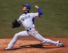 A man in a white baseball jersey with "KOREA" on the chest in blue and "17" on his leg in orange pitches a baseball from the pitcher's mound with his left hand. He is wearing a black baseball glove on his right hand.