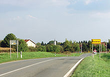 A view of a road from a side, showing two traffic lanes of a single carriageway as the road enters a village. A traffic sign indicating name of the village is visible on the right.