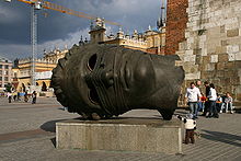 In an open plaza, a large, hollow head made of dark metal with empty eye sockets lies on its side on a concrete slab.