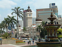 Street scene; a large fountain visible in the right foreground, a row of palm trees stretching away to the left, and in the centre of the image, across the street, a large white and beige stone building, with a tall domed central tower and two smaller domed towers to the right and left.