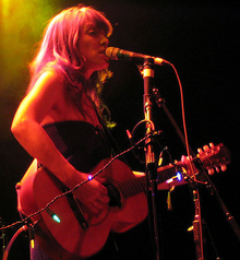 A female vocalist (wearing a strapless black dress) singing into a microphone on stage while playing guitar