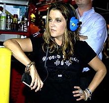 A woman in a black T shirt holds her left hand on her hip, while leaning against a chest-height surface with her right elbow. She is wearing blue headphones and looks towards her right.