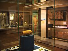View of the Cyrus Cylinder in its display cabinet, situated behind glass on a display stand. Other ancient Persian artifacts can be seen lining the room in the background.