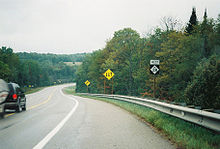A roadway curving down a slight incline and around a bend. On the side of the road are a series of sign posts with the M-28 reassurance marker, a yellow diamond with arrows indicating the traffic directions and a yellow diamond with an arrow warning of the curve in the road.