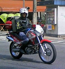 A firefighter in uniform, leather jacket and white helmet rides a motorcycle in red and white livery, equipped with emergency lights and a large pack of medical supplies, across a city street. Telephone booths and a newsstand are visible in the background.