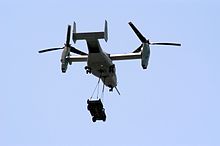  A MV-22 with its rotors up to vertical with a HMMWV vehicle hanging by two sling wires.