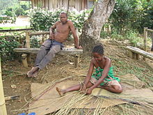 A Malagasy girls sits on the ground weaving with reeds, while an older gentleman sits on a bench above her.