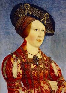 Woman in a heavily embroidered red dress wears an ornate hat with pearls and netting